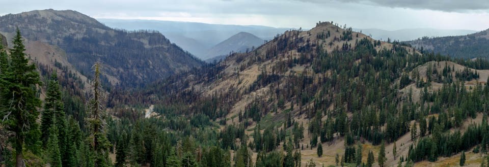 A panoramic photo of a valley in between volcanic peaks lined by conifer trees, a small number of which are brown from a recent wildfire.