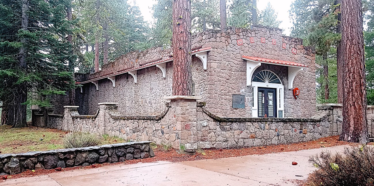 Historic stone and concrete building from the exterior. A stone and concrete half-wall surrounds the building. Steps lead to the front entrance double doors. Tall pine trees are amongst the building.