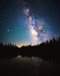 A dark sky filled with stars and a colorful cloud like streak rises above a black, tree-lined lake reflecting stars