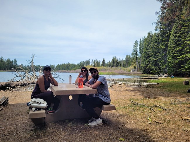 Two women and a man sit at a picnic bench next to a mountain lake.