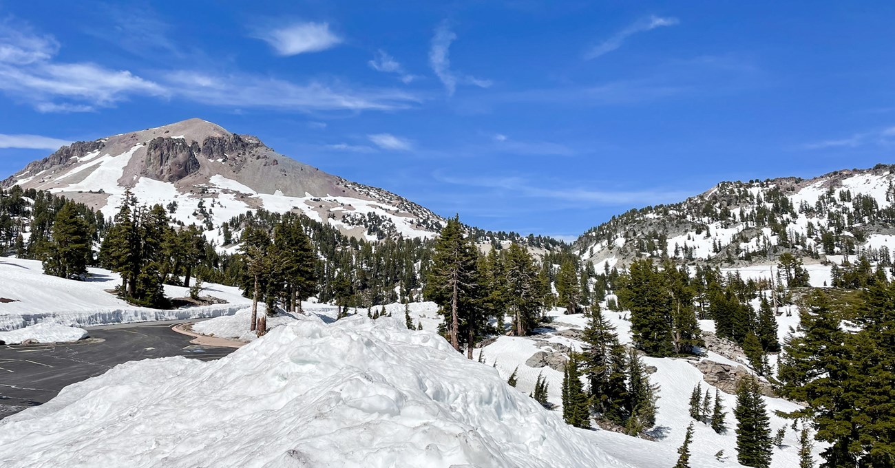 A panoramic photo of patchy snow on a volcanic, alpine landscape. A snow-free parking area at left rests below a large volcanic peak.