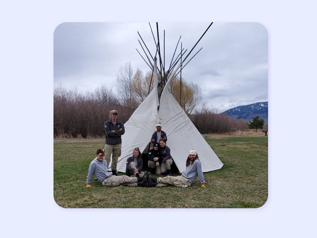 7 young people in AmeriCorps hoodies and jackets pose in front of teepee. Mountains behind.