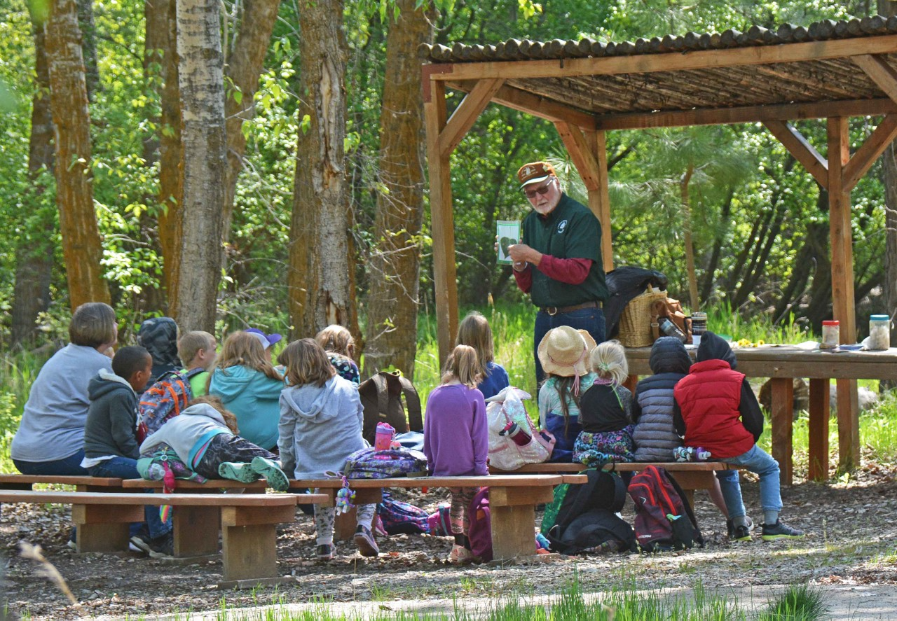 Ben Hagyard Travelers Rest 2022 Volunteer speaks to group of young students outside. Green trees and meadow behind. Volunteer is older man with white beard. He holds a photo of a leaf.
