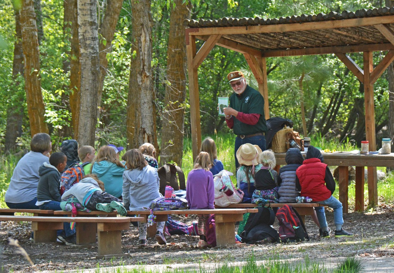 Volunteer speaks to group of young students outside. Green trees and meadow behind. Volunteer is older man with white beard. He holds a photo of a leaf.