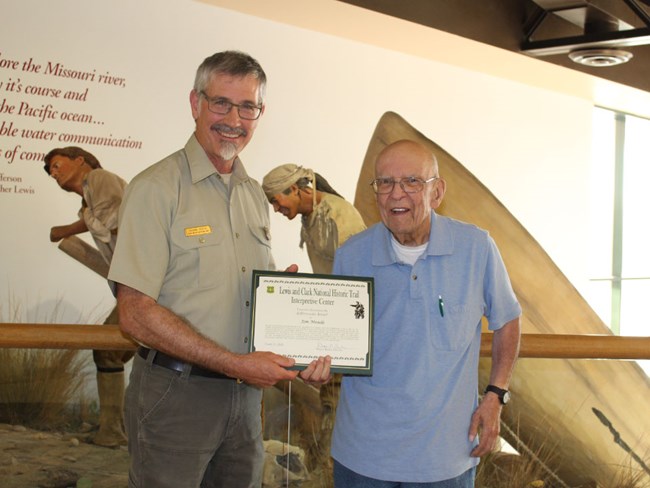 Man in Forest Service uniform poses with older man. They hold a certificate. Behind is a Lewis and Clark exhibit.