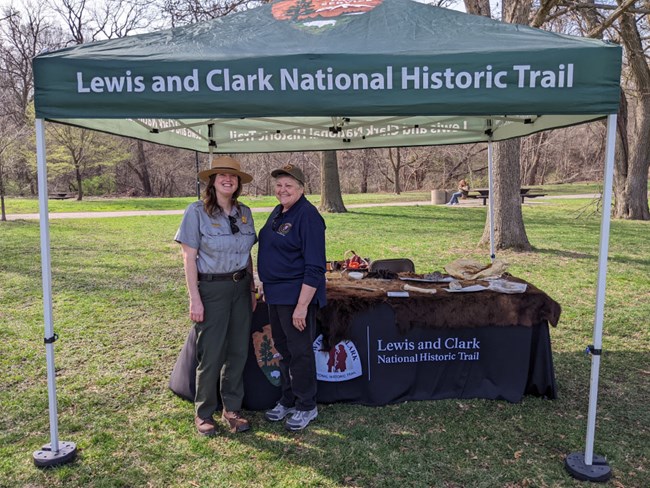 An NPS ranger and volunteer pose outside at a Lewis and Clark Trail table and tent