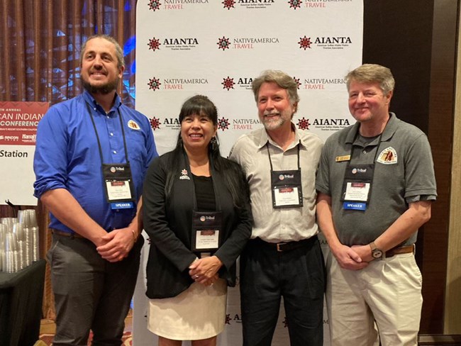 Four people pose. Backdrop reads AIANTA. Two men on left and right wear Lewis and Clark Trail polos. Woman and man in center.