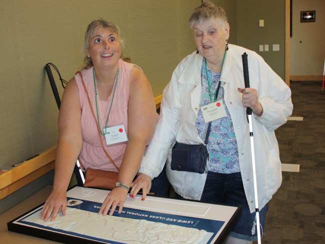 Two women with white canes smile as they feel a tactile map with braille captions.