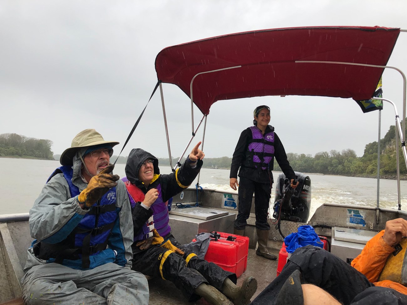 Young person manning engine of open boat in the rain. Two people point into the distance.