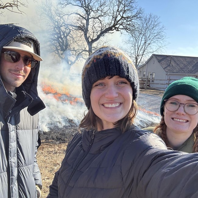 three people in winter clothing smile in front of a fire
