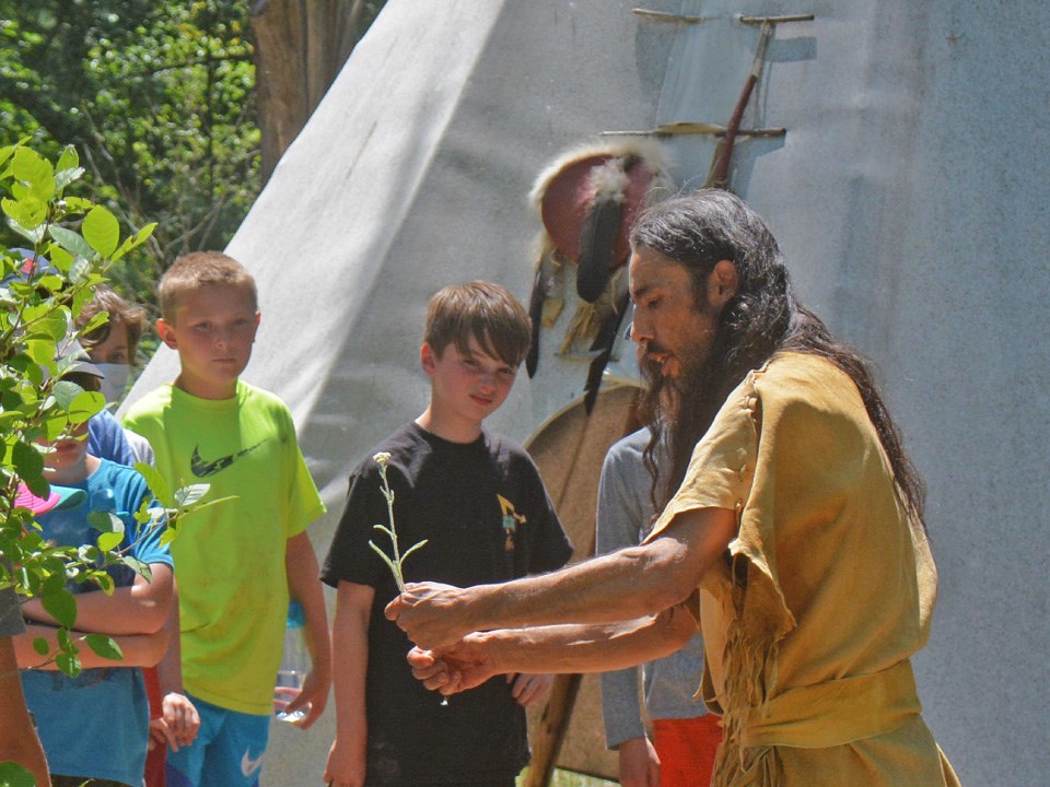 Person with long dark hair dressed in historic reenactment buckskins holds a plant and shows two young teens. Teepee behind.