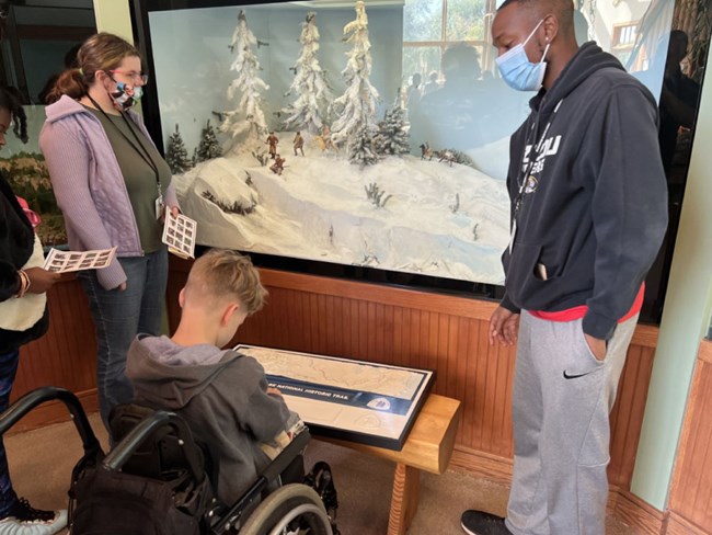 Child in wheelchair feels tactile map which is placed in front of him. Exhibits and two adults behind.