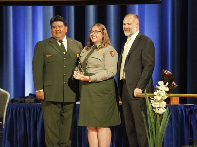 Three people in NPS uniform pose on a stage. Woman in center holds glass award.