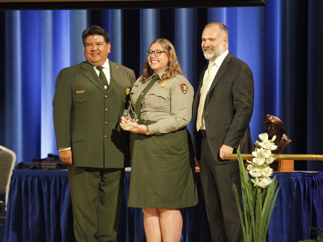 Freeman-Tidlen-Award-Sams-Campbell-Beaudreau Three people in NPS uniform pose on a stage. Woman in center holds glass award.