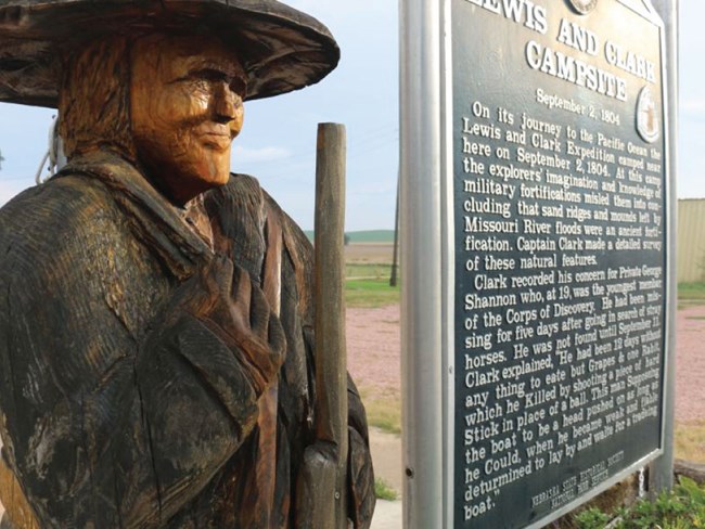 Wooden carved statue of explorer with wide brimmed hat. Lewis and Clark Trail sign.