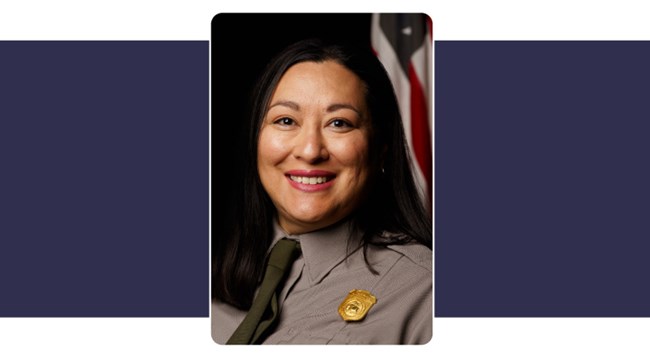 Woman wearing blue Lewis and Clark button up shirt long dark hair smiles. American Flag behind.