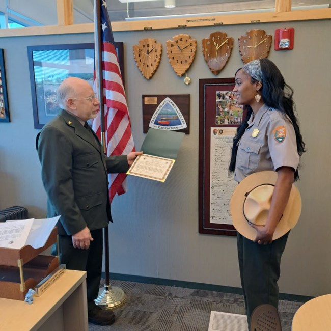 Two National Park Service rangers face each other. A man hands a young woman a certificate.