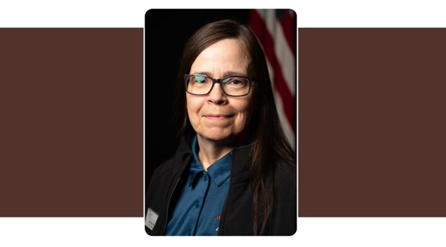 Portrait of Woman wearing button down shirt and jacket. Long dark hair glasses. American Flag background.
