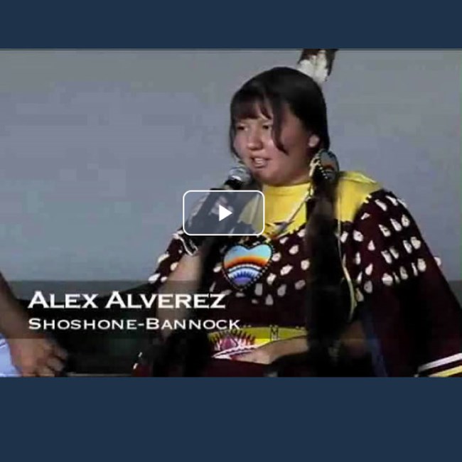 Screenshot of video. Native woman in regali hold microphone. Caption reads Alex Alveraz, Shoshone-Bannock
