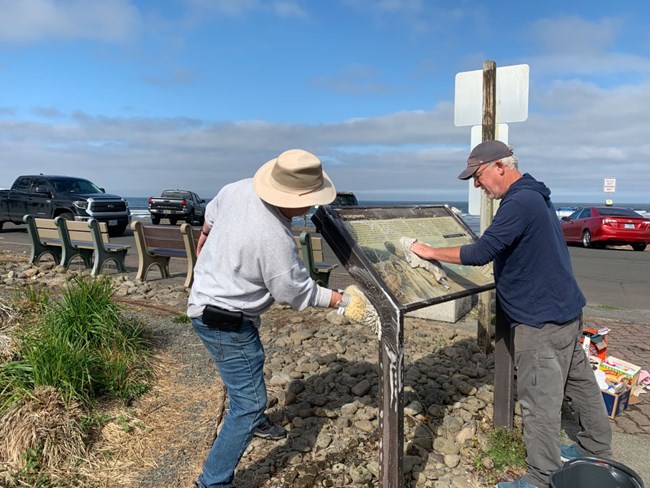 Two people work on installing a wayside. Behind them is a parking lot at a Pacific Coast seaside viewpoint.