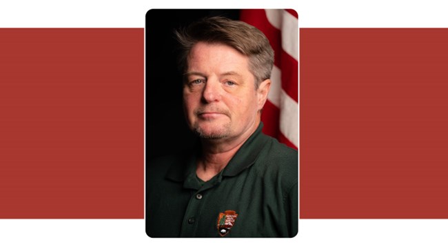 portrait of man with short brown hair. American flag in background.