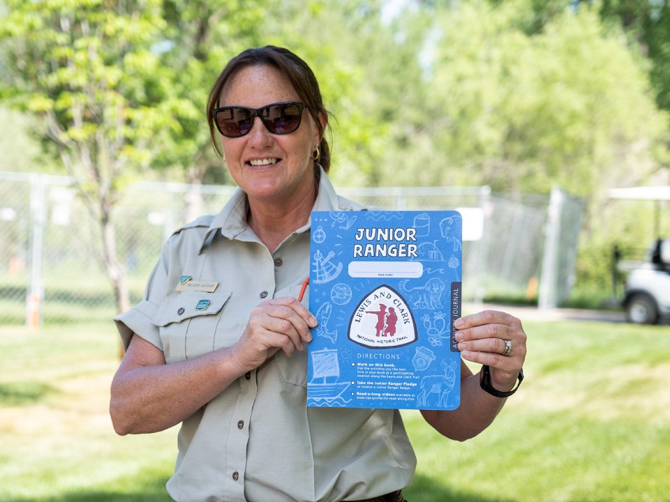 Ranger with sunglasses and pony tail smiles and holds up blue activity booklet. Booklet reads Junior Ranger. Lewis and Clark Trail logo.