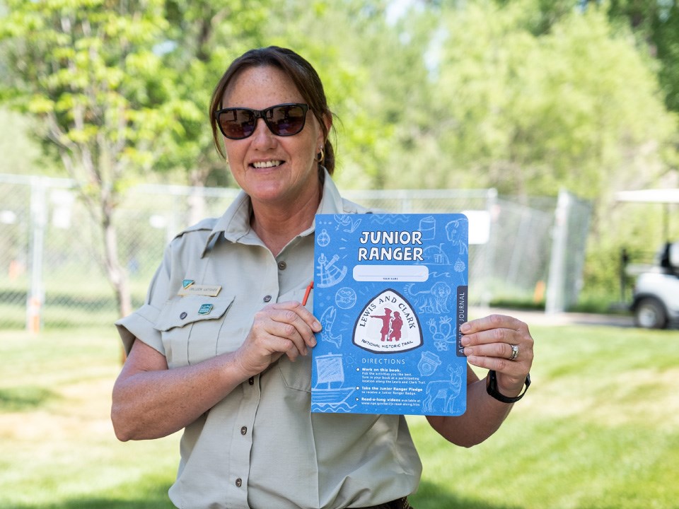 Lewis-and-Clark-Trail-Junior-RAnger-BLM-Ranger-Colleen-Pompeys-Pillar-National-Monument-2021 Ranger with sunglasses and pony tail smiles and holds up blue activity booklet. Booklet reads Junior Ranger. Lewis and Clark Trail logo.
