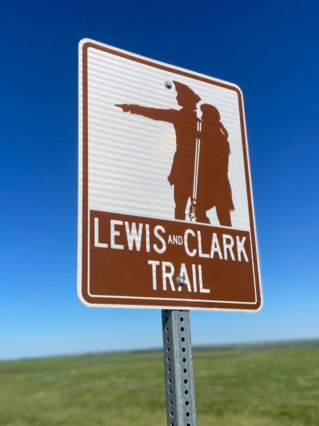 Photo of Lewis and Clark Trail Sign. Two male explorers point west. Bright blue sky, green prairie beyond.