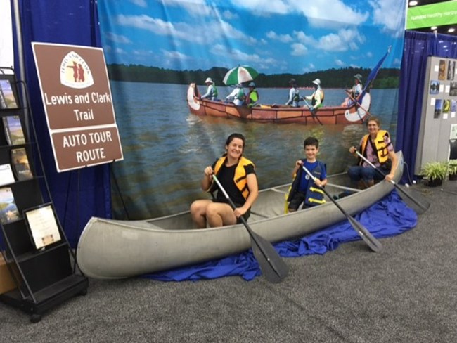 Family poses in canoe display at a fair. Exhibit features a Lewis and Clark Trail sign and a photo backdrop of paddlers on a river.