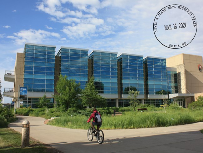 Person bikes past large office building. Circular stamp reads Lewis & Clark Natl Historic Trail. Omaha, NE. March 16, 2021