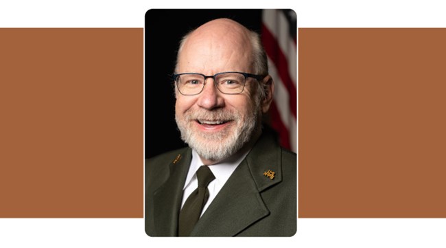 portrait of man with white beard and glasses in formal National Park Service Uniform tie and jacket. American flag background.