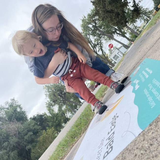 Woman holds toddler as the child walks on a sidewalk sticker