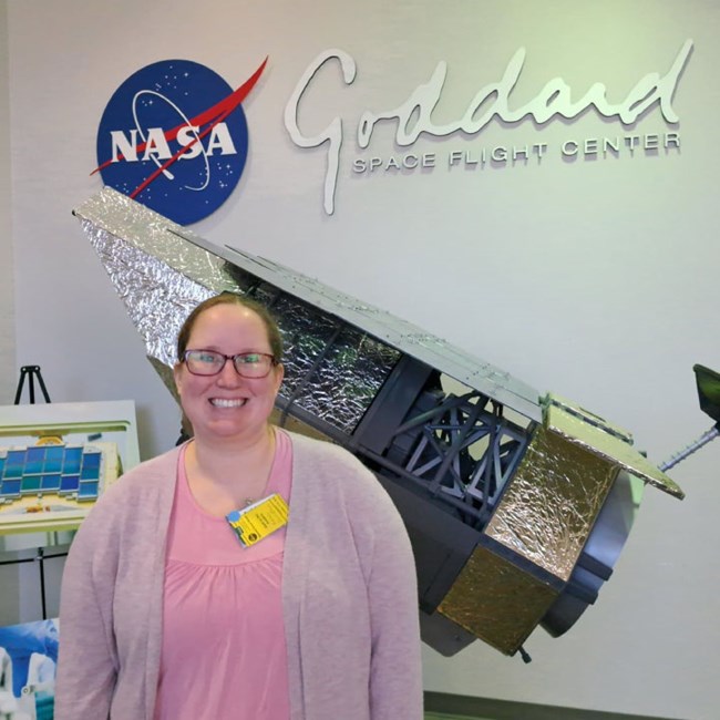 Woman poses in front of NASA Goddard Space Center sign. Display of rocket behind.