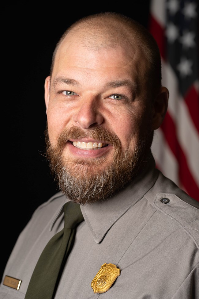 a man in a park ranger uniform smiles in front of the American flag