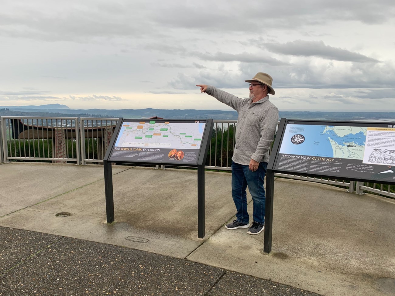 Man poses with two new Lewis and Clark Trail waysides. 1st sign is a map of US. 2nd is a map of Columbia River. Evergreen hills behind.