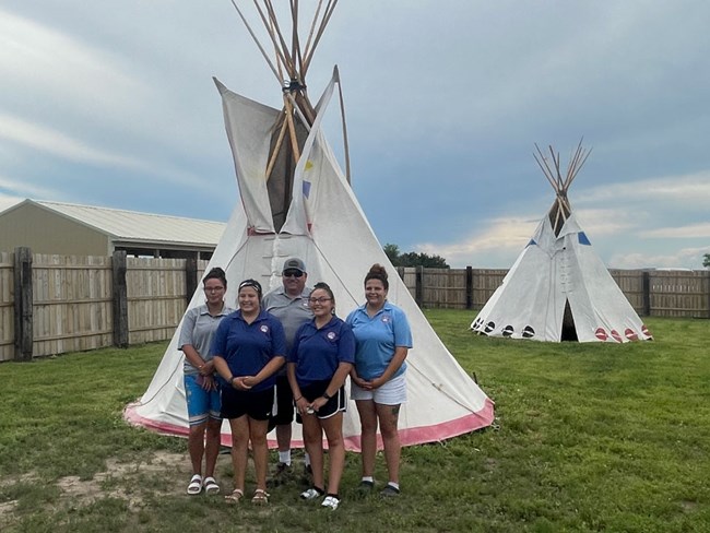 5 people pose in front of a teepee. Second teepee behind.