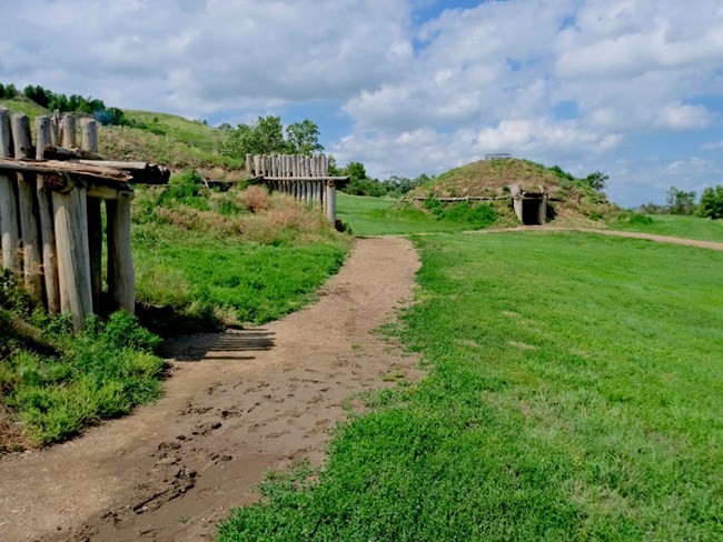 earth lodge village. Wooden entries protrude from 3 green grass covered earth lodges. A path winds through. Blue sky.