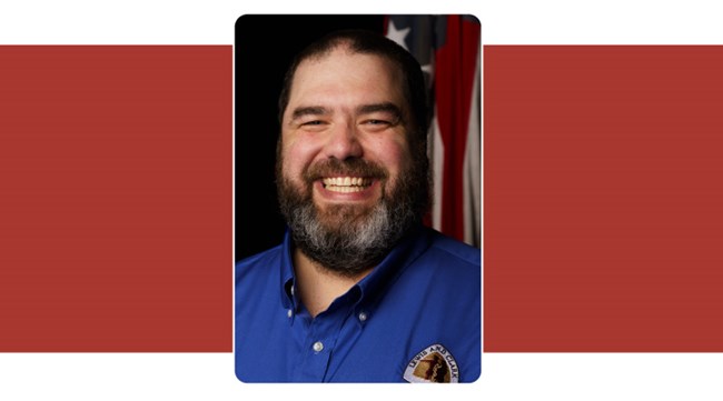 portrait of man with dark brown beard, smiling. American Flag background