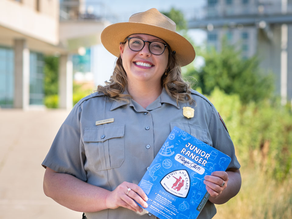 Campbell Tilden Award Junior Ranger Park ranger in uniform smiles and holds blue junior ranger booklet with Lewis and Clark Trail logo.