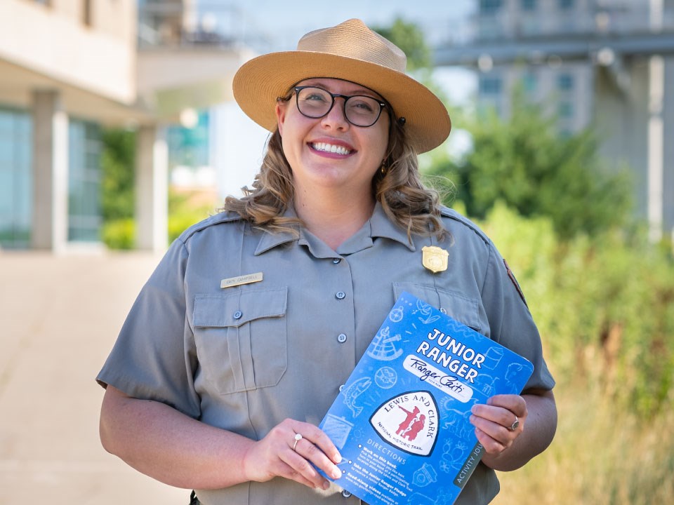 Park ranger in uniform smiles and holds blue junior ranger booklet with Lewis and Clark Trail logo.