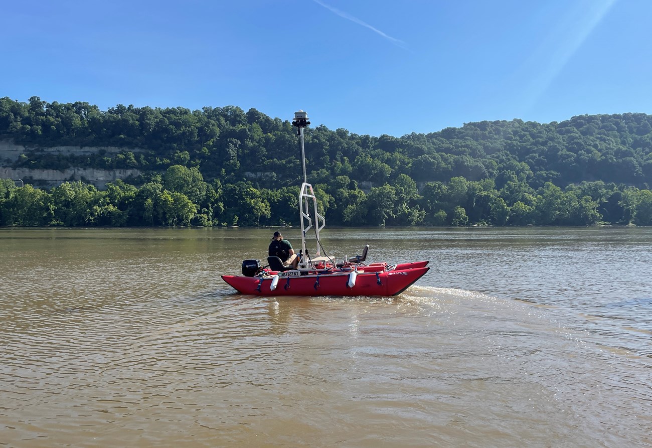 Red rubber double hulled boat with camera mounted on a mast along a tree lined river.