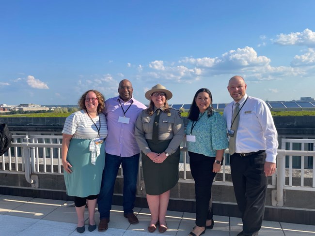 5 people pose on rooftop with city background. Women in center wear NPS uniform. Four others, two women and two men wear business casual.