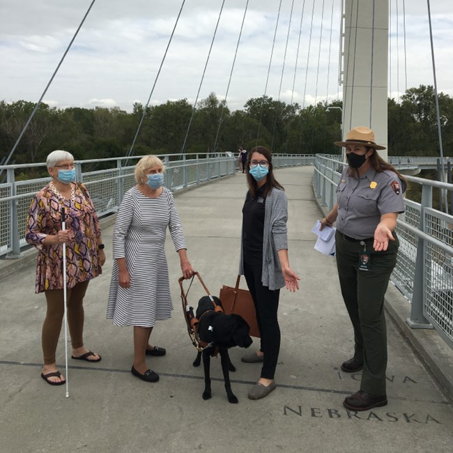 Four people pose on pedestrian bridge over line that reads Nebraska. Iowa. One woman holds white walking stick. One woman holds leash of black seeing eye dog. Park ranger in uniform. All wear masks and gesture happily with arms out.