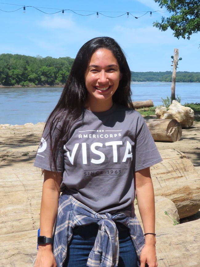 Young person smiles and stands of riverbank. Dark hair past shoulders. Shirt reads VISTA
