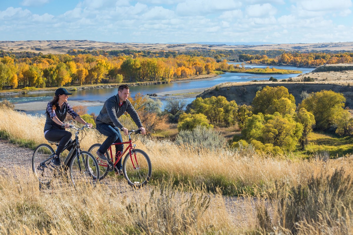 Two people ride bikes along a path. Mountains, trees in autumn gold, and a small river in background.
