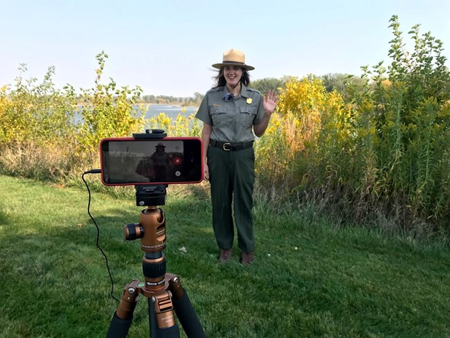 Ranger stands along riverbank and waves. A tripod holds up a phone which records her.