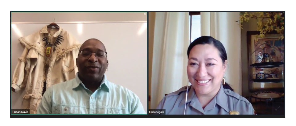 Screenshot of two people on a video call. Man on left wears a light blue shirt. A replica early 1800s explorer outfit hangs behind. Woman in park ranger uniform smiles.