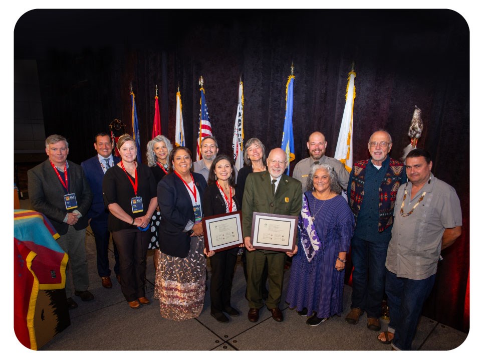 Honoring-Tribal-Legacies-NIEA-group-photo 2021 13 people pose for a photo. Some in uniform, one with a large necklace, one with a tribal print vest, they appear to be from varied background.