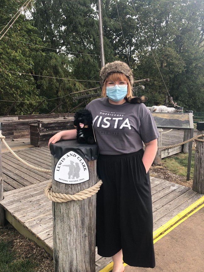 Young person stands next to Lewis and Clark Trail Sign. Shirt reads VISTA. Keelboat behind.