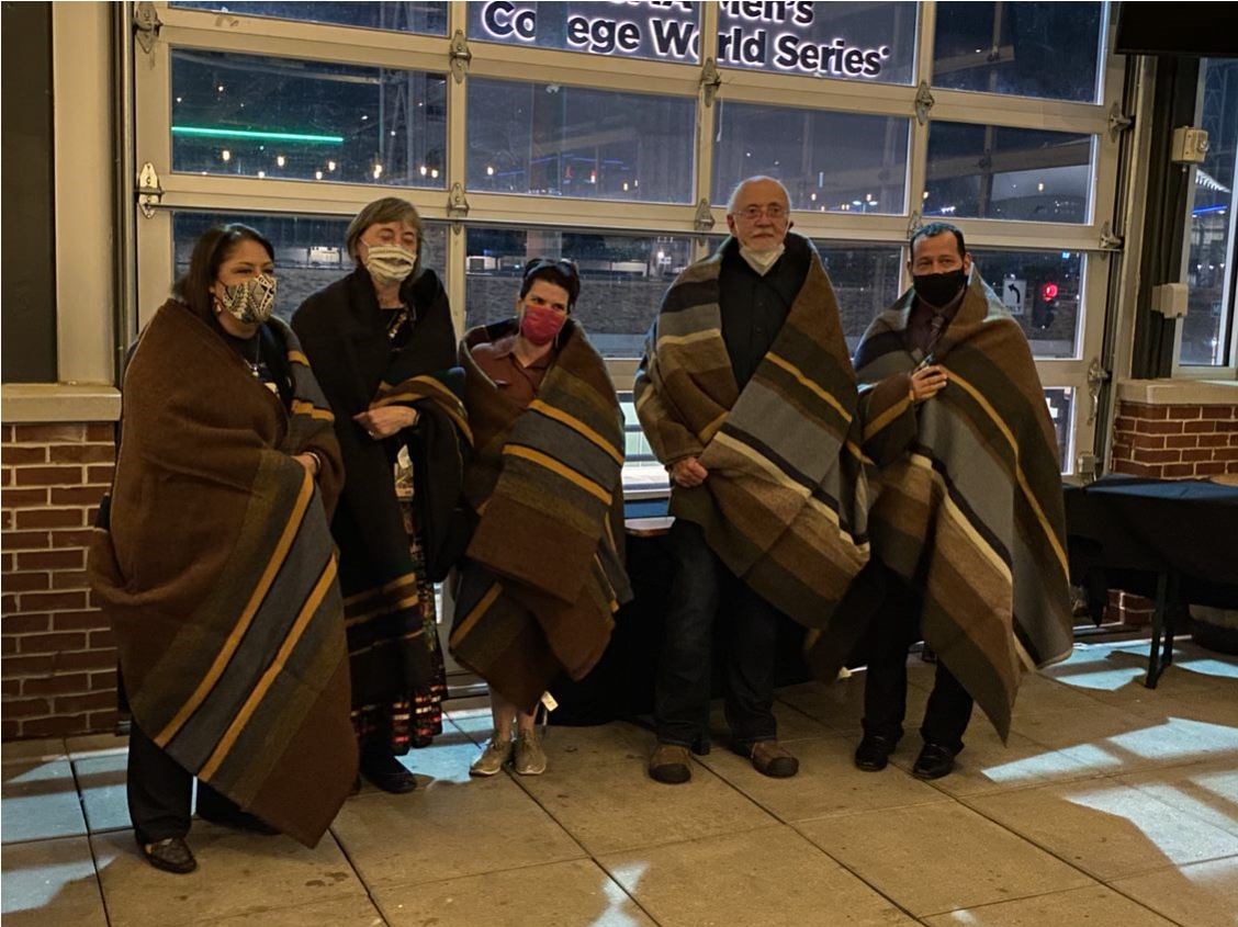 A group of five people pose with large wool blankets pulled over their shoulders. They wear masks. City in background.